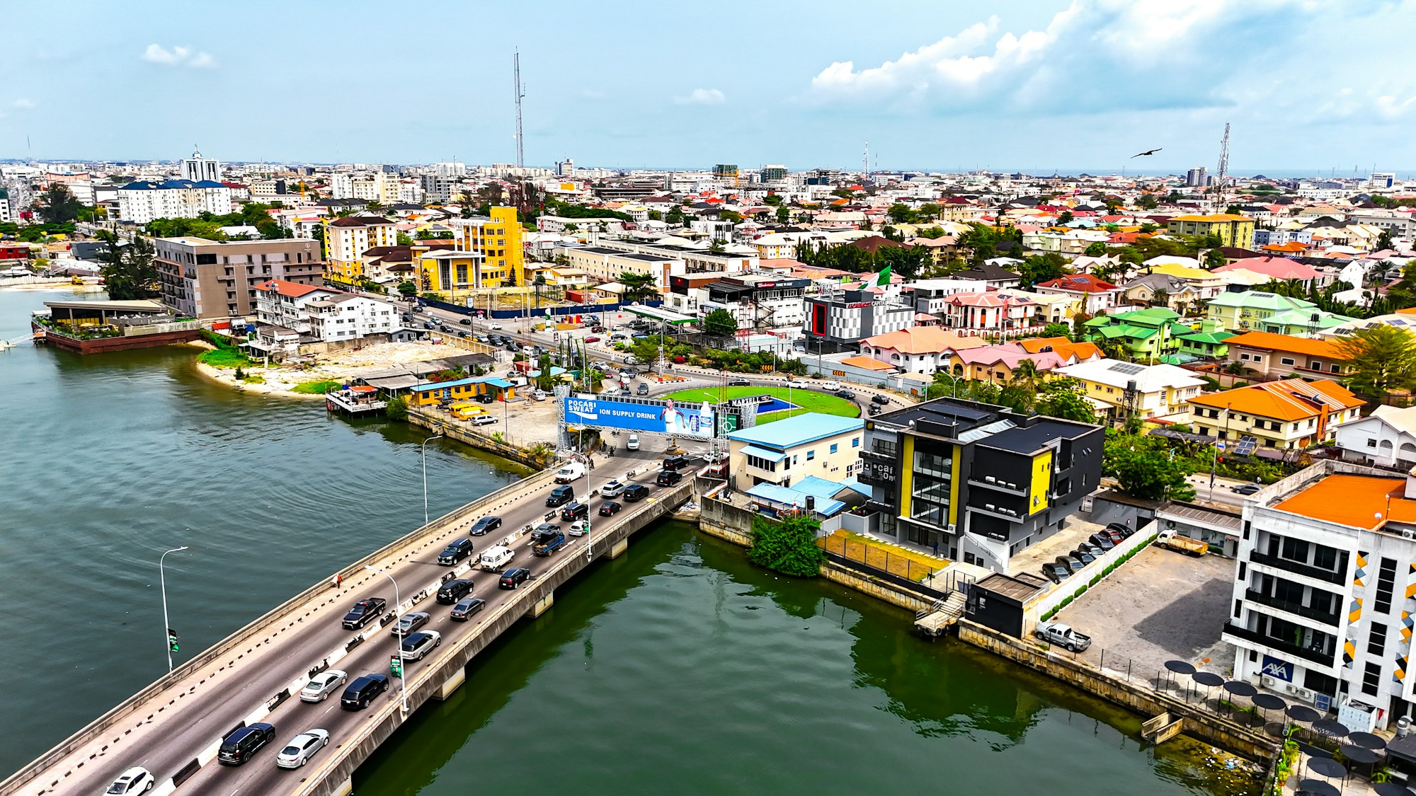 Vue aérienne d’une ville côtière d’Afrique de l’Ouest : pont routier chargé, lagune et quartiers denses.